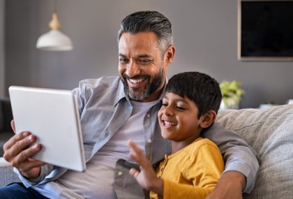 A father and son, sitting on a couch, laughing while watching something on a tablet.