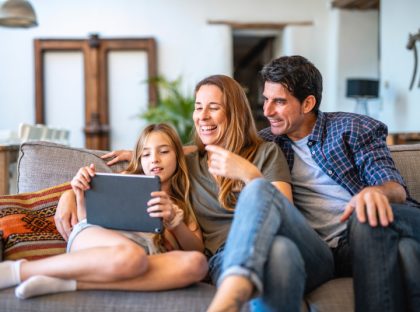 A mother, father and daughter are sitting on a couch.They are smiling and watching the tablet that the daughter is holding.